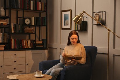 Photo of Young woman reading book in armchair indoors. Home library