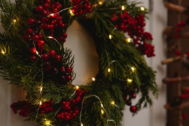 Beautiful Christmas wreath with red berries and fairy lights hanging on white door, closeup