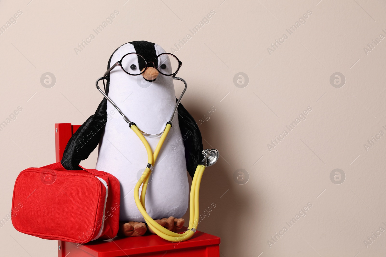 Photo of Toy penguin with eyeglasses, stethoscope and first aid bag on beige background, space for text. Pediatrician practice