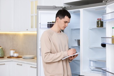 Photo of Man writing notes near empty refrigerator in kitchen