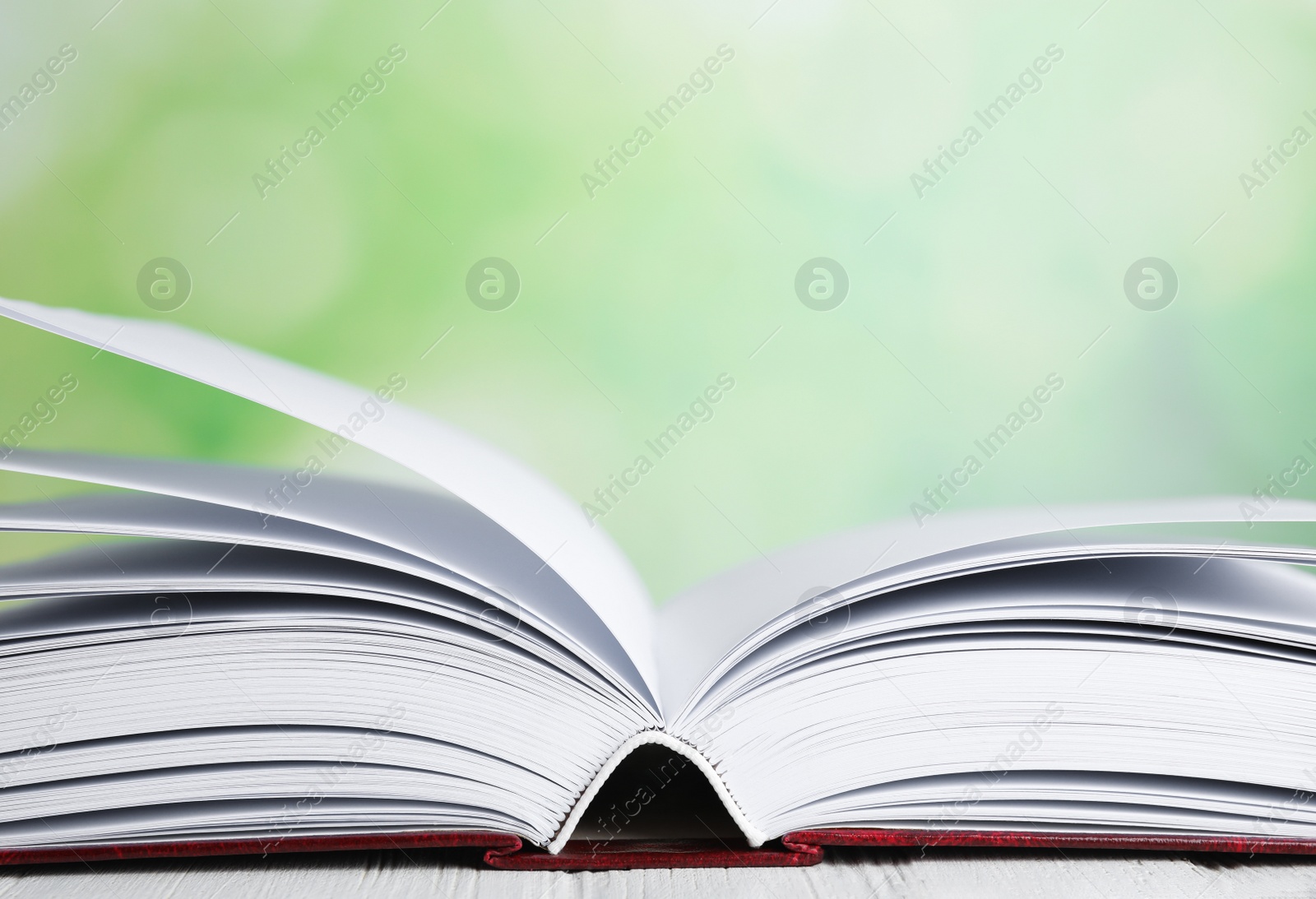 Photo of Open book on white wooden table against blurred green background, closeup. Space for text