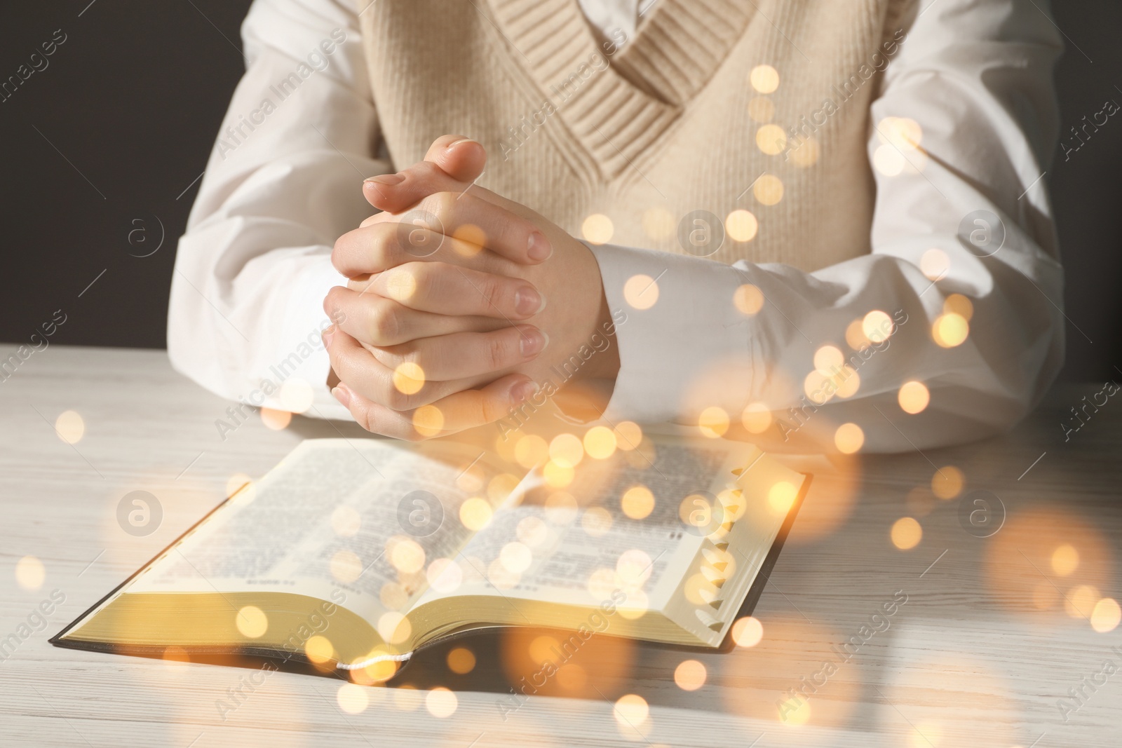 Image of Woman praying over Bible at white wooden table, closeup. Bokeh effect