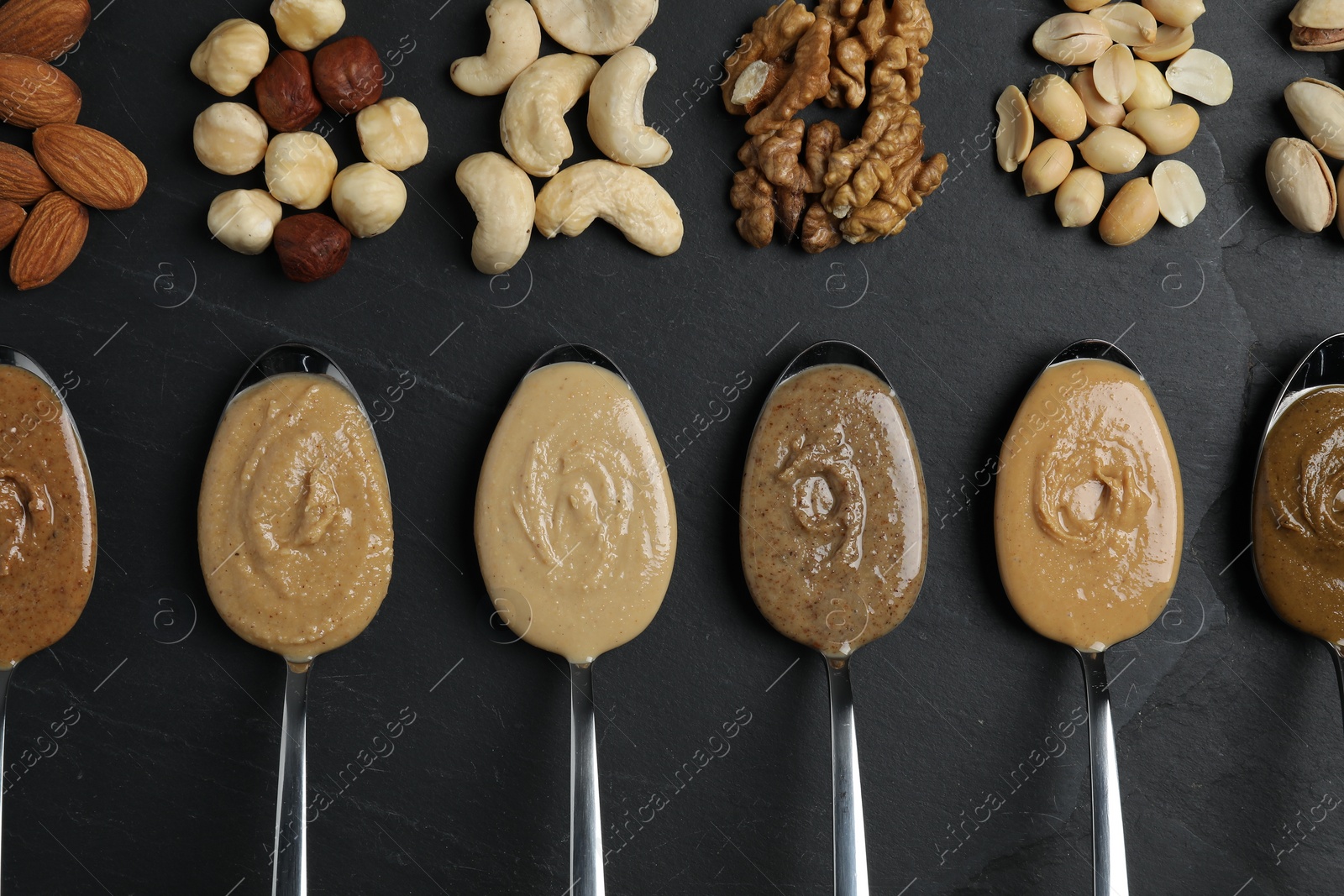 Photo of Tasty nut butters in spoons and raw nuts on dark gray textured table, flat lay