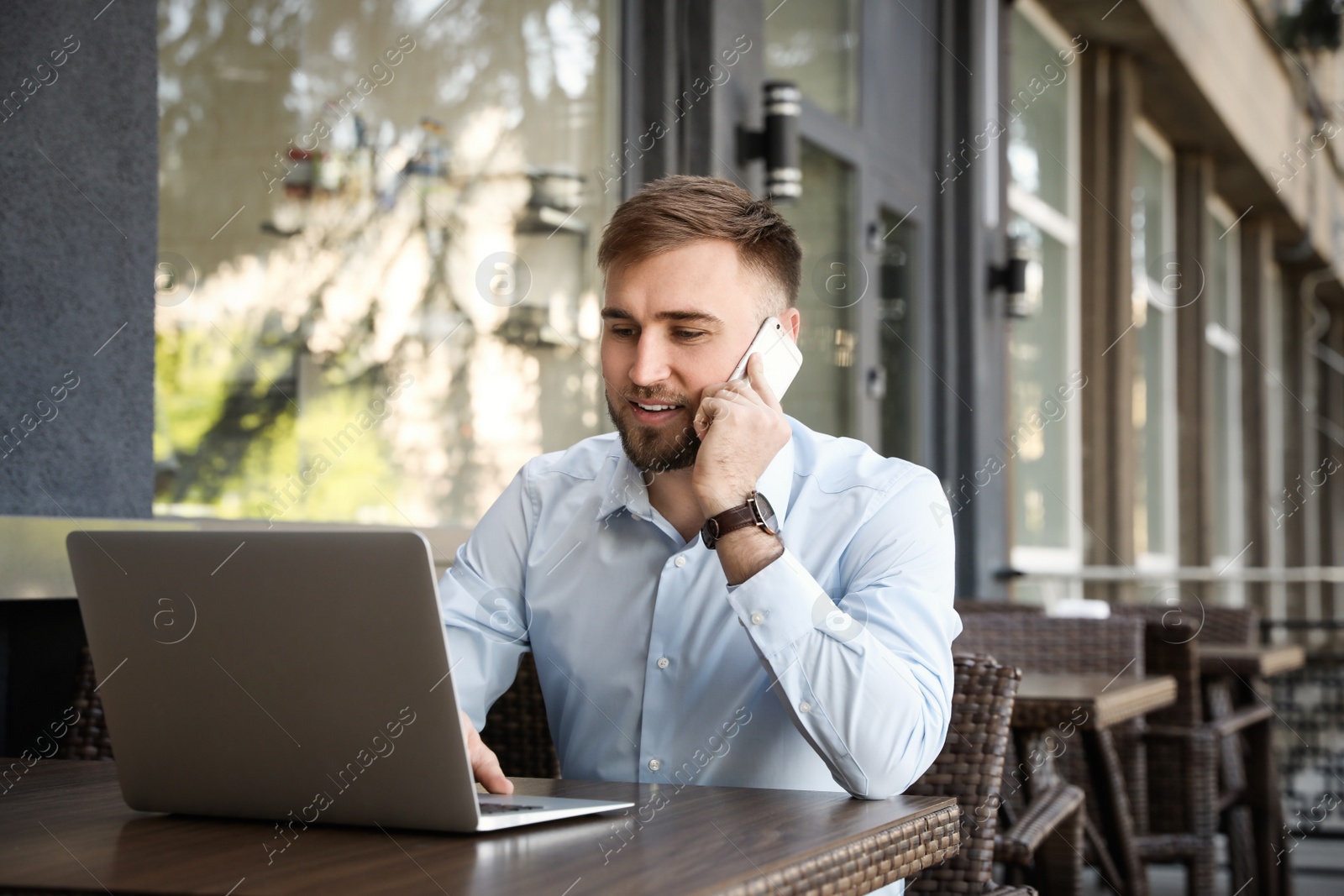 Image of Happy young man talking on phone while using laptop at outdoor cafe