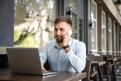 Image of Happy young man talking on phone while using laptop at outdoor cafe