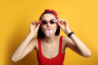 Photo of Fashionable young woman in pin up outfit blowing bubblegum on yellow background