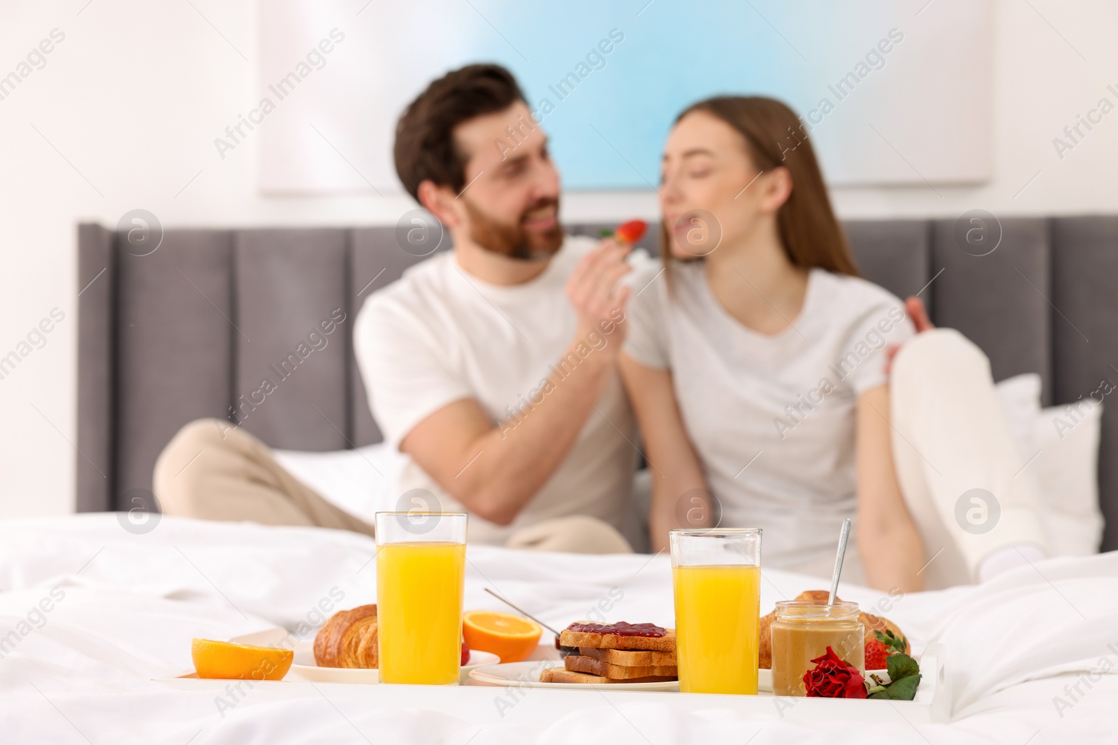 Photo of Tray with tasty breakfast on bed. Husband feeding his wife in bedroom, selective focus