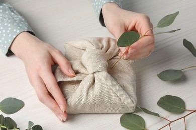 Photo of Furoshiki technique. Woman decorating gift wrapped in fabric with eucalyptus branch at white wooden table, closeup