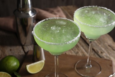 Delicious Margarita cocktail in glasses, limes and shaker on wooden table