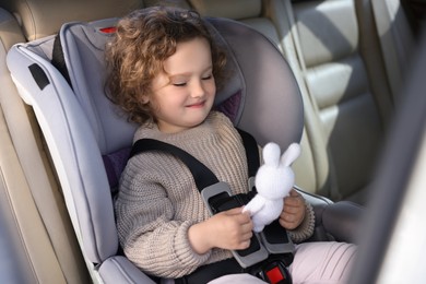 Photo of Cute little girl with toy rabbit sitting in child safety seat inside car