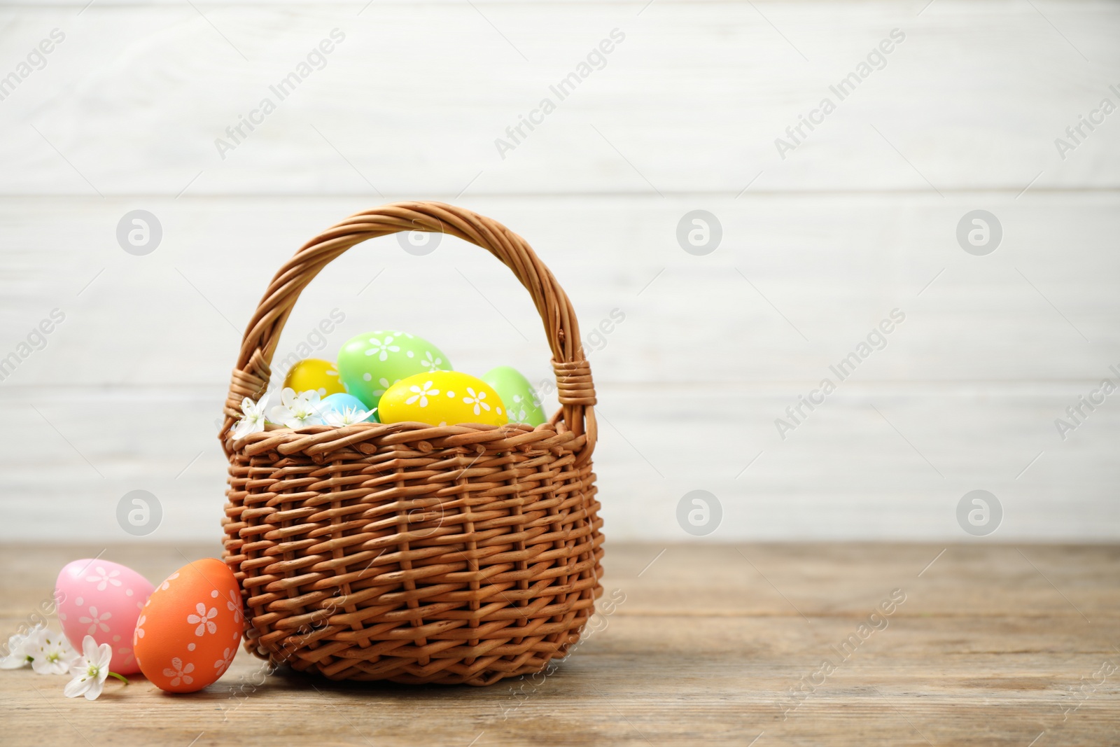 Photo of Colorful Easter eggs in basket and flowers on wooden table. Space for text