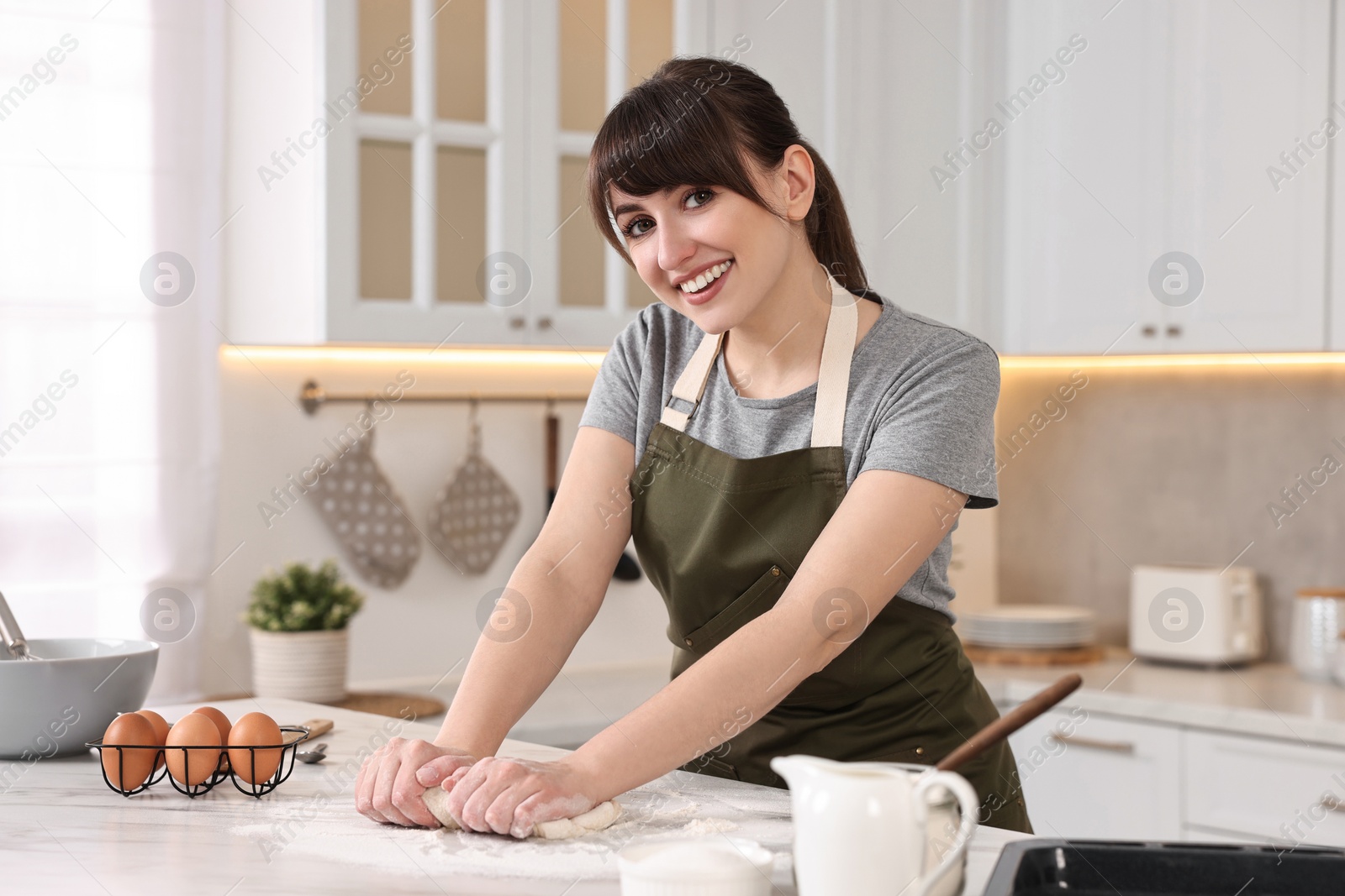 Photo of Happy young housewife kneading dough at white marble table in kitchen