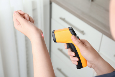 Woman measuring temperature with non contact infrared thermometer indoors, closeup