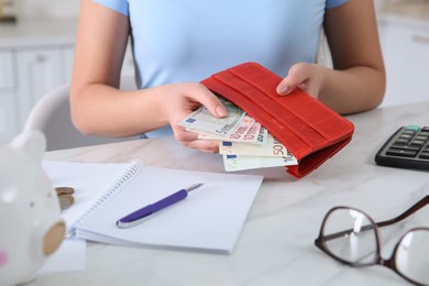 Photo of Young woman putting money into wallet at table in kitchen, closeup