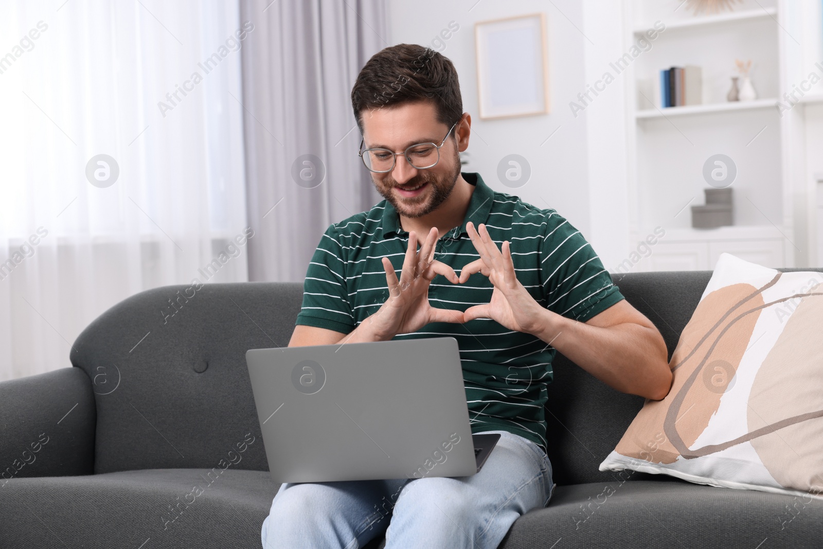 Photo of Happy man making heart with hands during video chat via laptop at home. Long-distance relationship