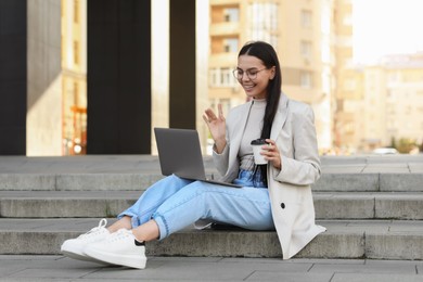 Happy young woman using modern laptop for video call on stairs outdoors