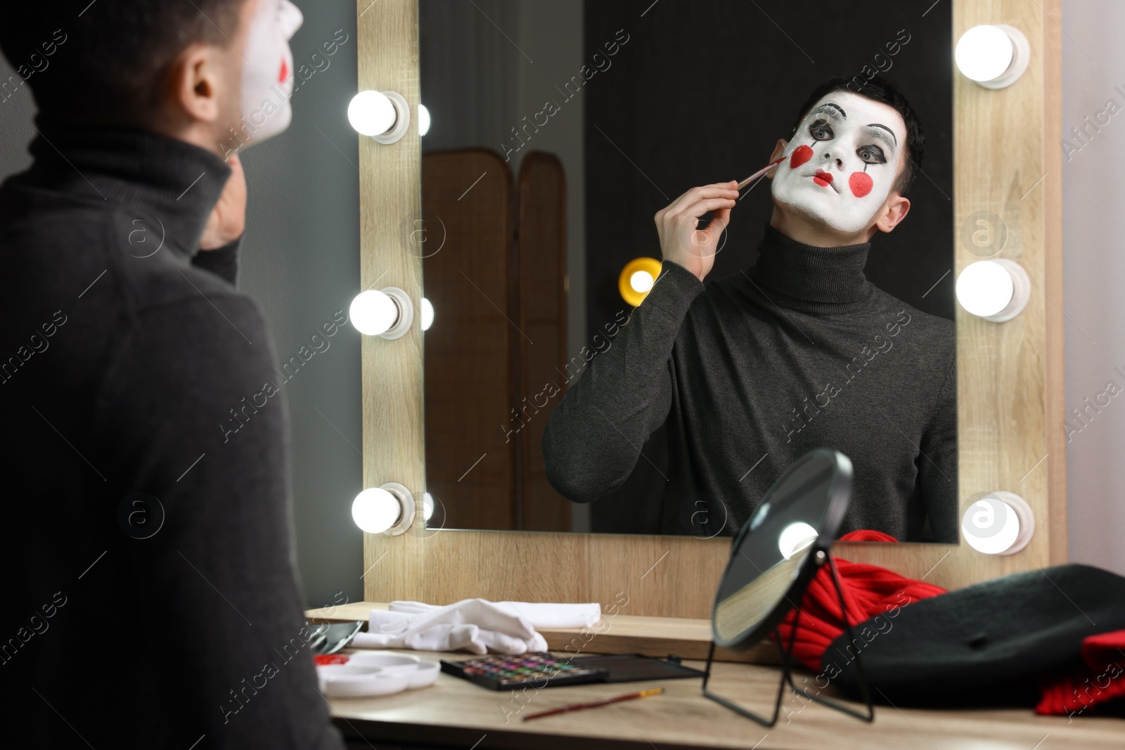 Photo of Young man applying mime makeup near mirror in dressing room