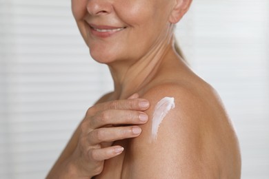 Happy woman applying body cream onto shoulder on light background, closeup