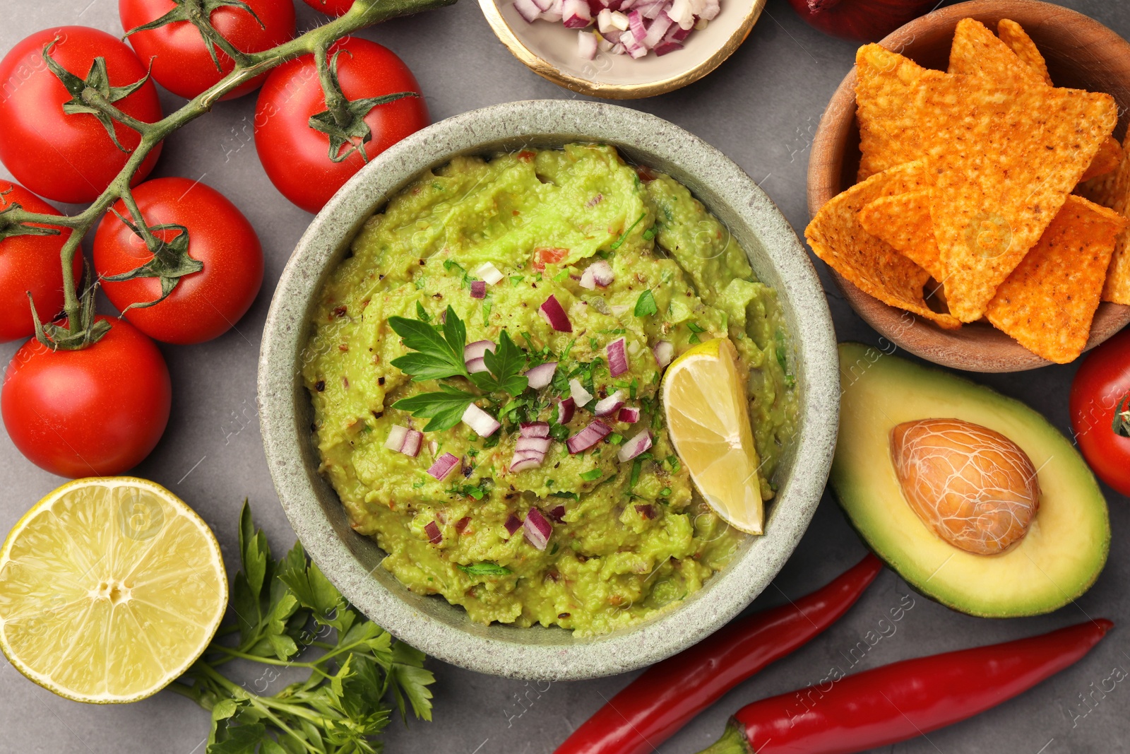 Photo of Bowl of delicious guacamole, nachos chips and ingredients on grey table, flat lay