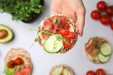 Photo of Woman holding crunchy buckwheat cakes with prosciutto, pieces of tomato and cucumber slice over white table, closeup