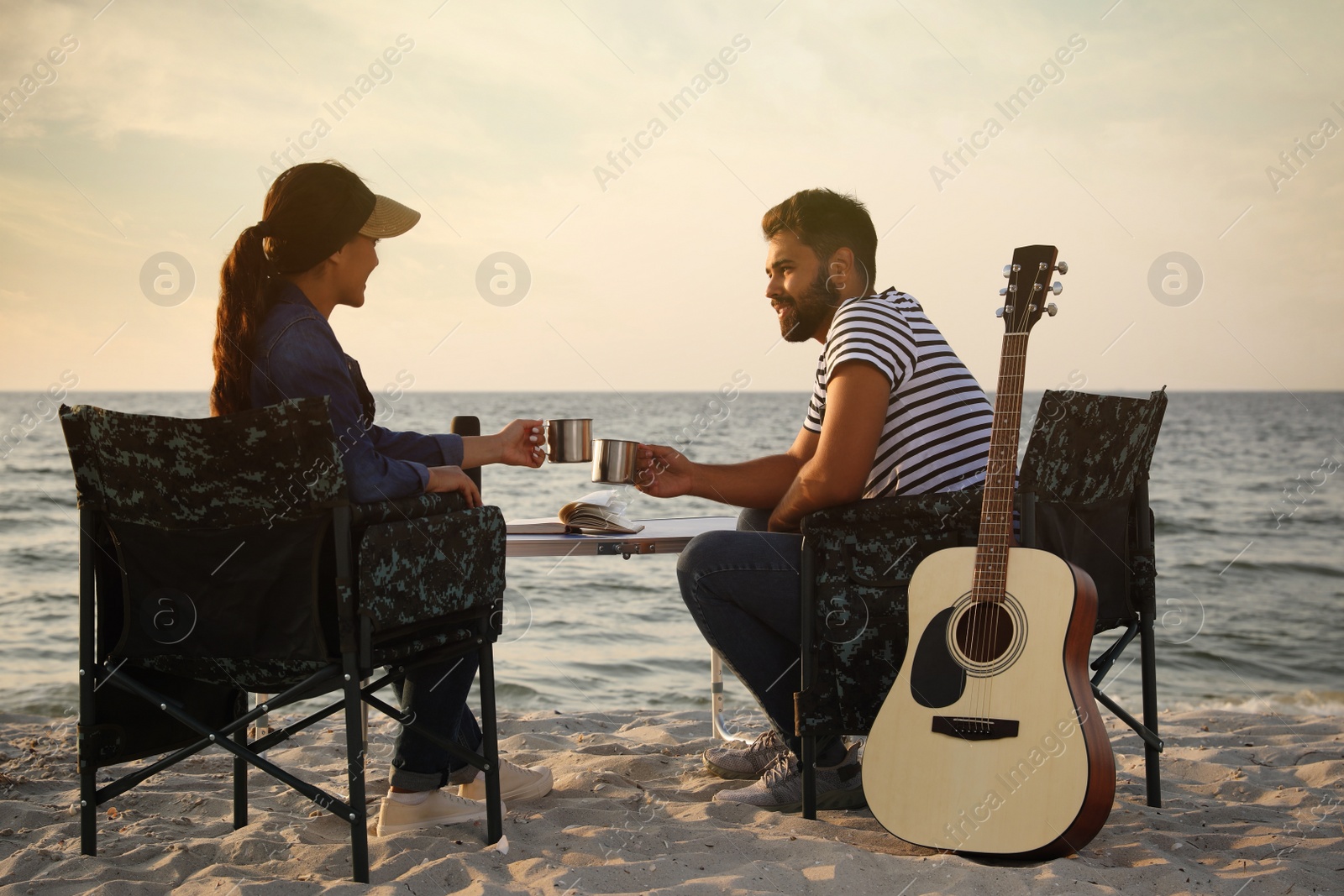 Photo of Couple sitting in camping chairs and clinking mugs on beach