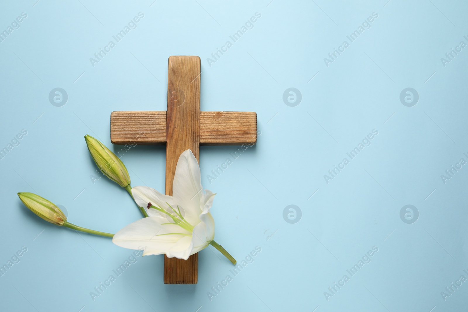 Photo of Wooden cross and lily flowers on light blue background, top view with space for text. Easter attributes