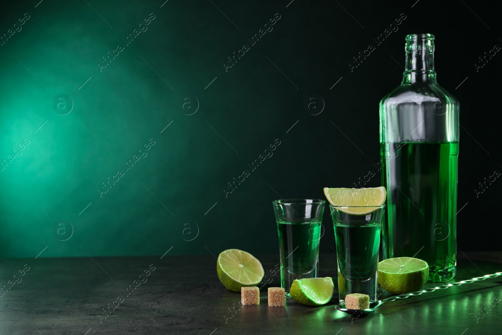 Photo of Absinthe in shot glasses, spoon, brown sugar cubes and lime on gray textured table against green background, space for text. Alcoholic drink