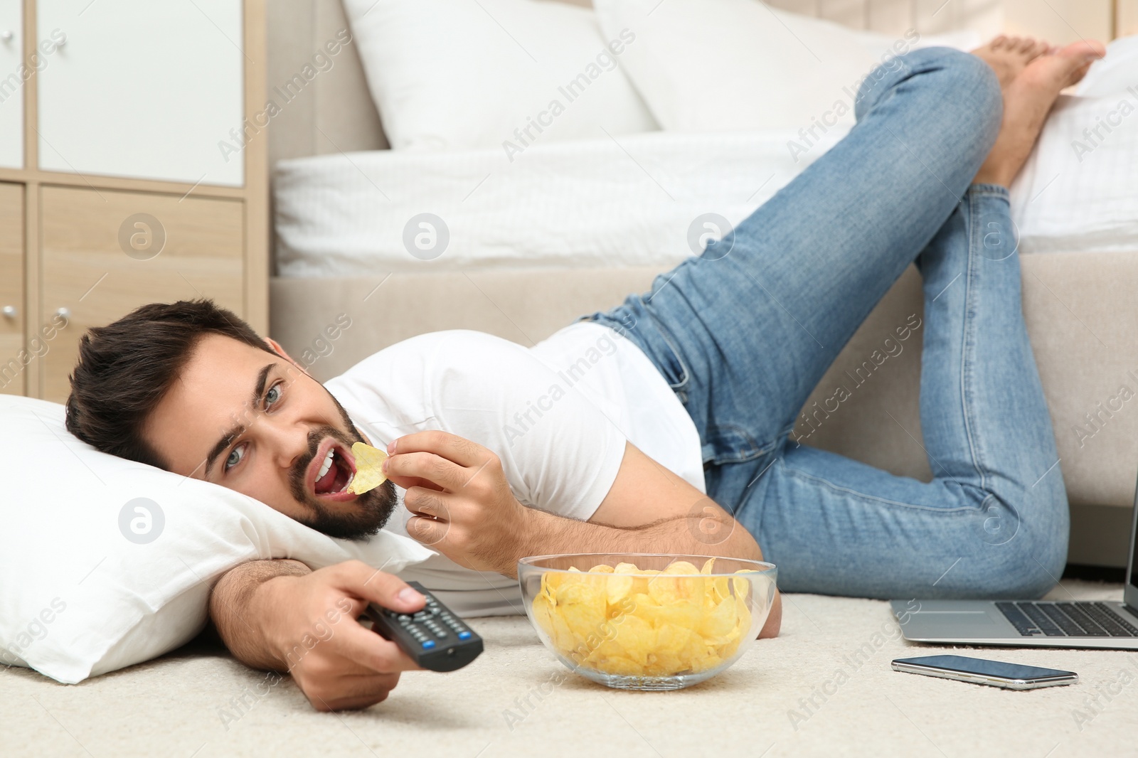 Photo of Lazy young man with bowl of chips watching TV while lying on floor at home