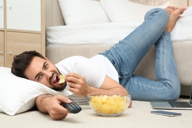 Lazy young man with bowl of chips watching TV while lying on floor at home