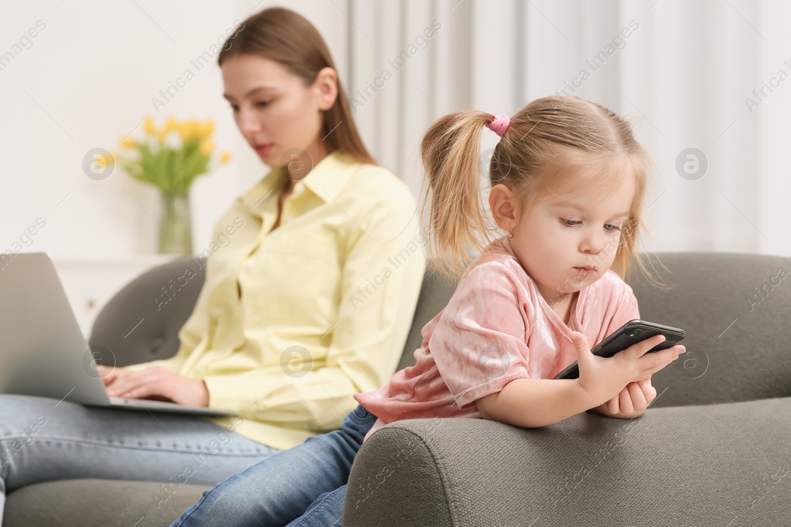 Photo of Mother working remotely on laptop while her daughter playing with smartphone at home, selective focus