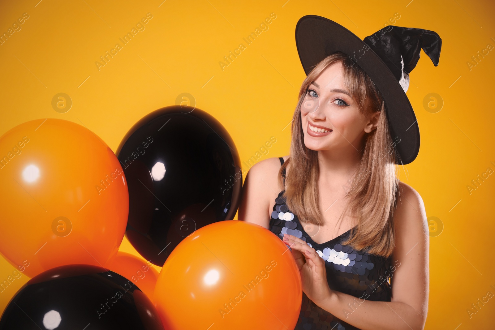 Photo of Beautiful woman in witch costume with balloons on yellow background. Halloween party