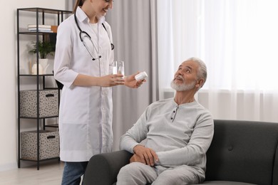 Photo of Young healthcare worker giving bottle of pills and water to senior man indoors