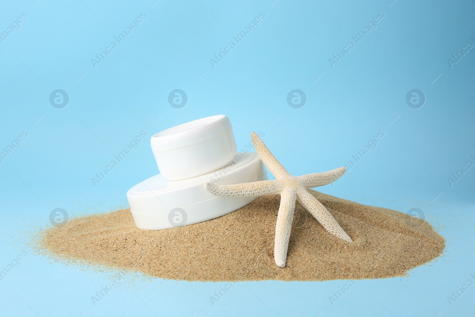 Photo of Jars with cream and starfish on sand against light blue background. Cosmetic products