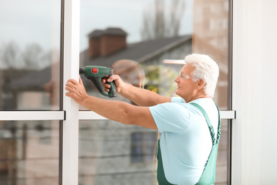 Photo of Mature construction worker repairing plastic window with electric screwdriver indoors