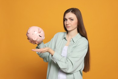 Sad young woman with piggy bank on orange background
