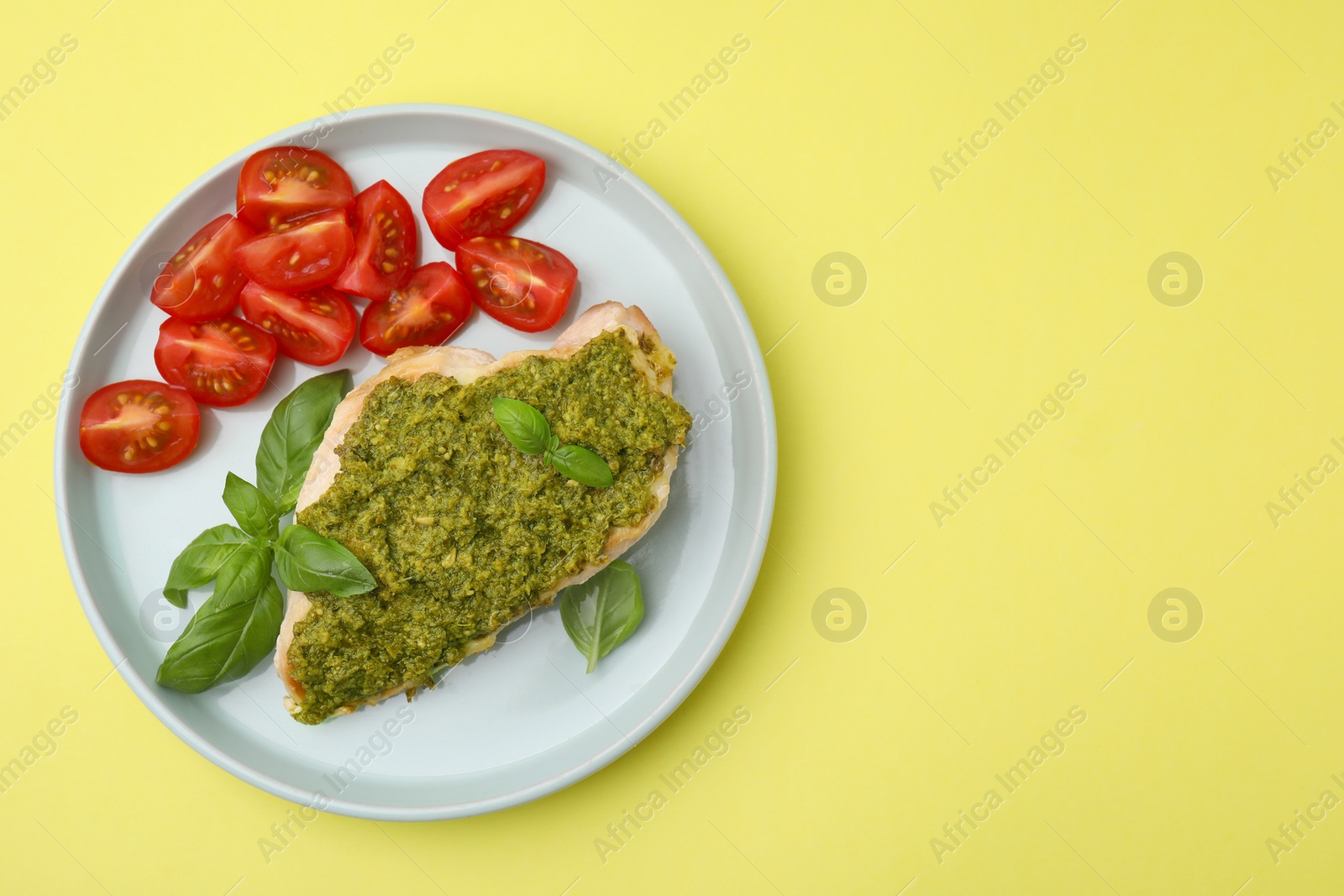 Photo of Delicious chicken breast with pesto sauce, tomatoes and basil on yellow table, top view. Space for text