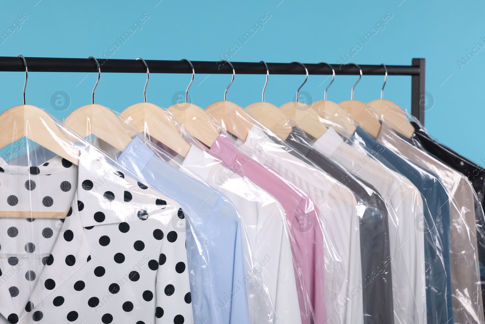 Photo of Dry-cleaning service. Many different clothes in plastic bags hanging on rack against light blue background