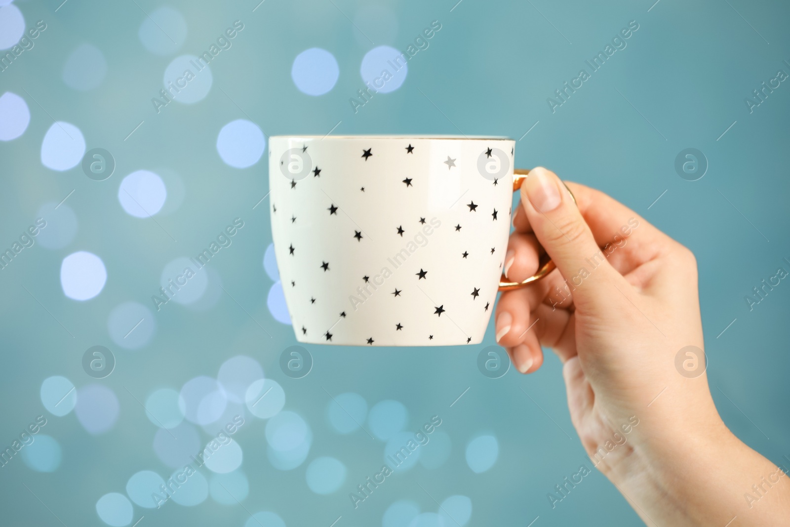 Photo of Woman holding cup of tasty hot drink against light blue background with blurred lights, closeup. Bokeh effect