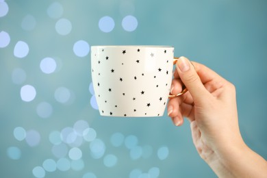 Photo of Woman holding cup of tasty hot drink against light blue background with blurred lights, closeup. Bokeh effect