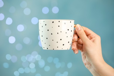 Woman holding cup of tasty hot drink against light blue background with blurred lights, closeup. Bokeh effect