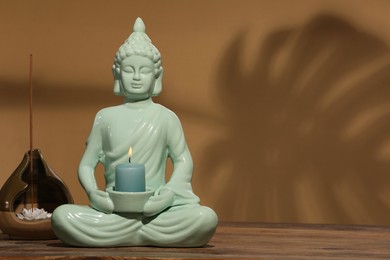 Photo of Buddhism religion. Decorative Buddha statue with burning candle and incense stick on wooden table against light brown wall, space for text