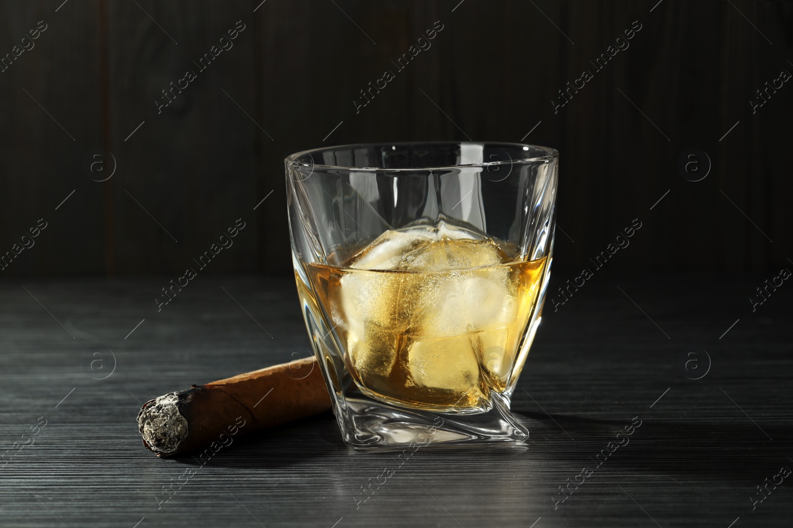 Photo of Glass of whiskey with ice cubes and smoldering cigar on black wooden table, closeup