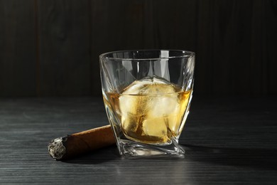 Photo of Glass of whiskey with ice cubes and smoldering cigar on black wooden table, closeup