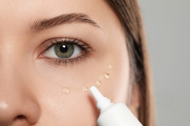 Young woman applying cream under eye on light grey background, closeup