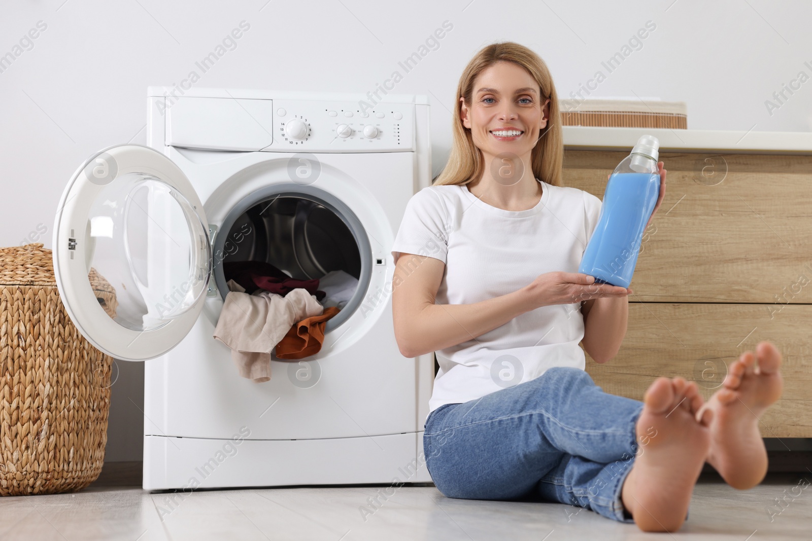 Photo of Woman sitting on floor near washing machine and holding fabric softener in bathroom, space for text