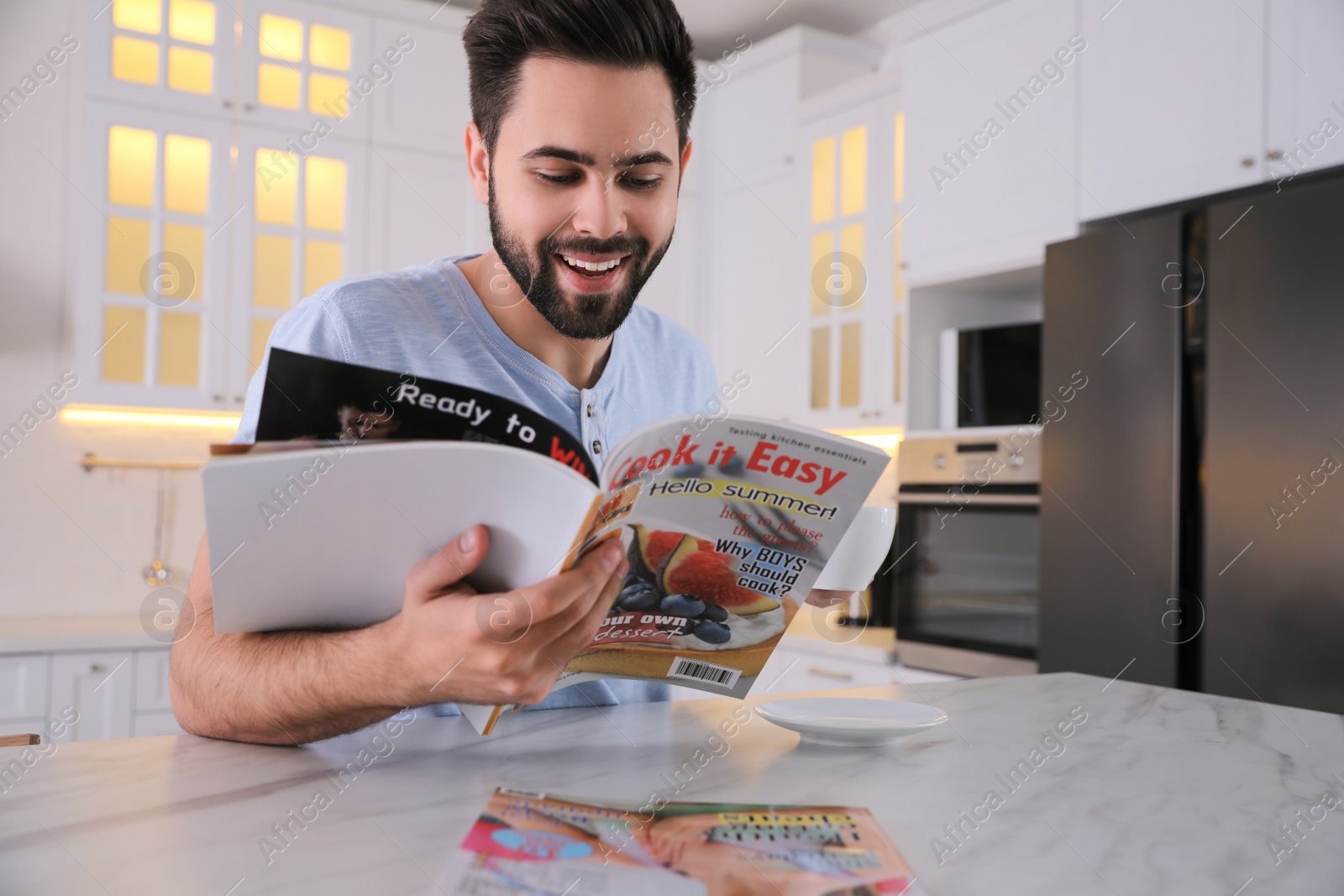 Photo of Young man with cup of drink reading magazine at table in kitchen