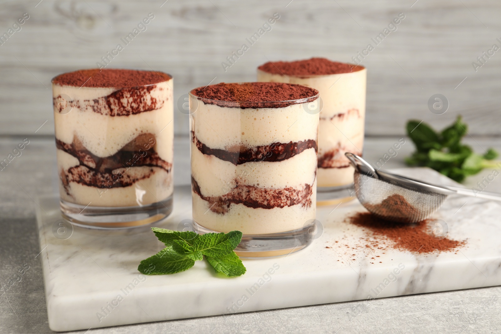 Photo of Delicious tiramisu in glasses, mint and sieve with cocoa powder on light textured table, closeup