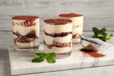 Delicious tiramisu in glasses, mint and sieve with cocoa powder on light textured table, closeup