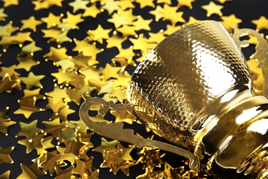 Photo of Gold trophy cup and confetti on black background, closeup. Space for text