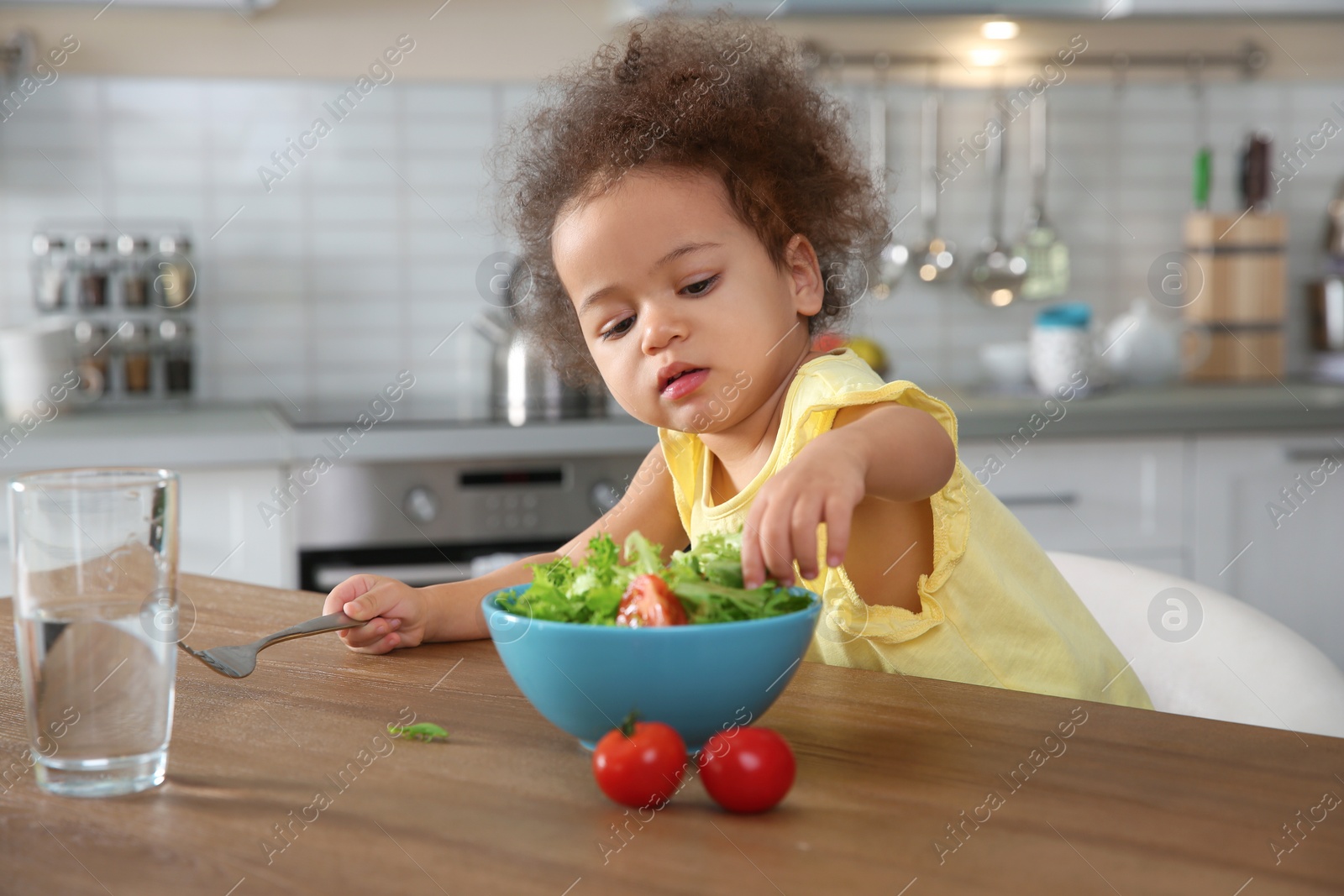 Photo of Cute African-American girl eating vegetable salad at table in kitchen